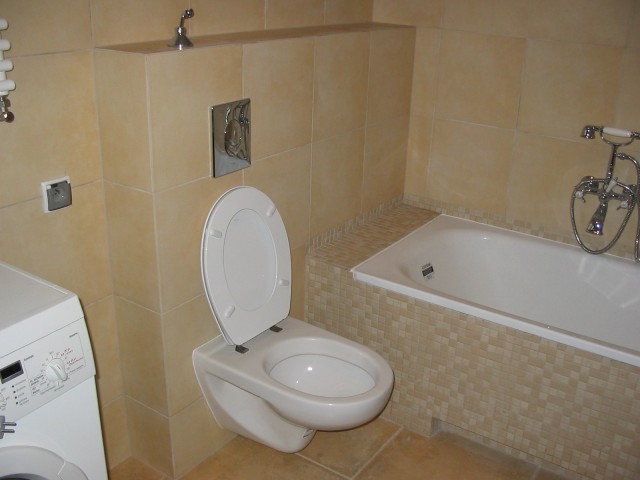 Jasna łazienka z beżowymi płytkami, wisząca toaleta z otwartą klapą, wanna obudowana mozaiką i pralka z przodu, chromowana bateria wannowa.