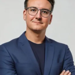 Portret mężczyzny w okularach i granatowym garniturze, z zegarkiem na ręce i skrzyżowanymi ramionami, uśmiechniętego na jasnym tle.