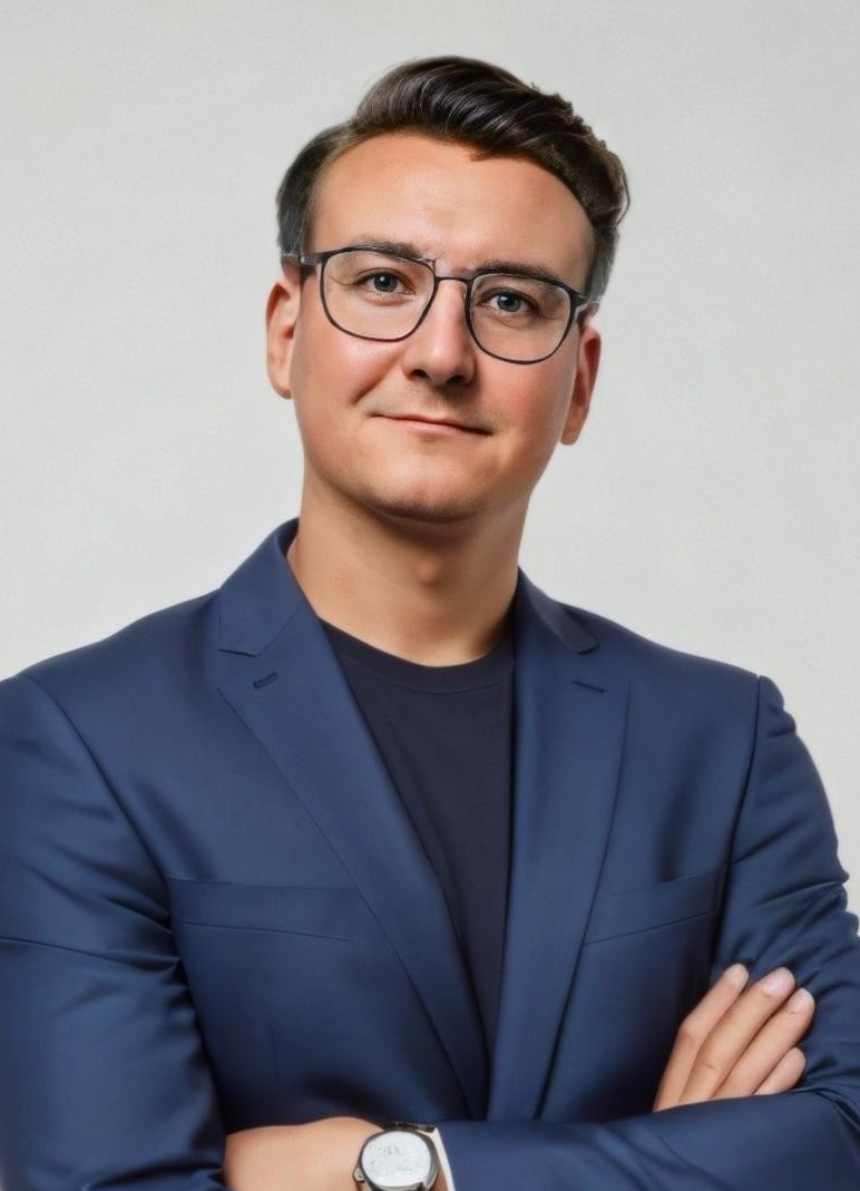 Portret mężczyzny w okularach i granatowym garniturze, z zegarkiem na ręce i skrzyżowanymi ramionami, uśmiechniętego na jasnym tle.