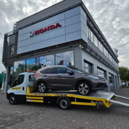 Brązowa Honda SUV na ż&oacute;łtej lawecie przed salonem Hondy. Pomoc drogowa 24h, usługi transportowe. Samochody na parkingu w tle, pochmurne niebo.