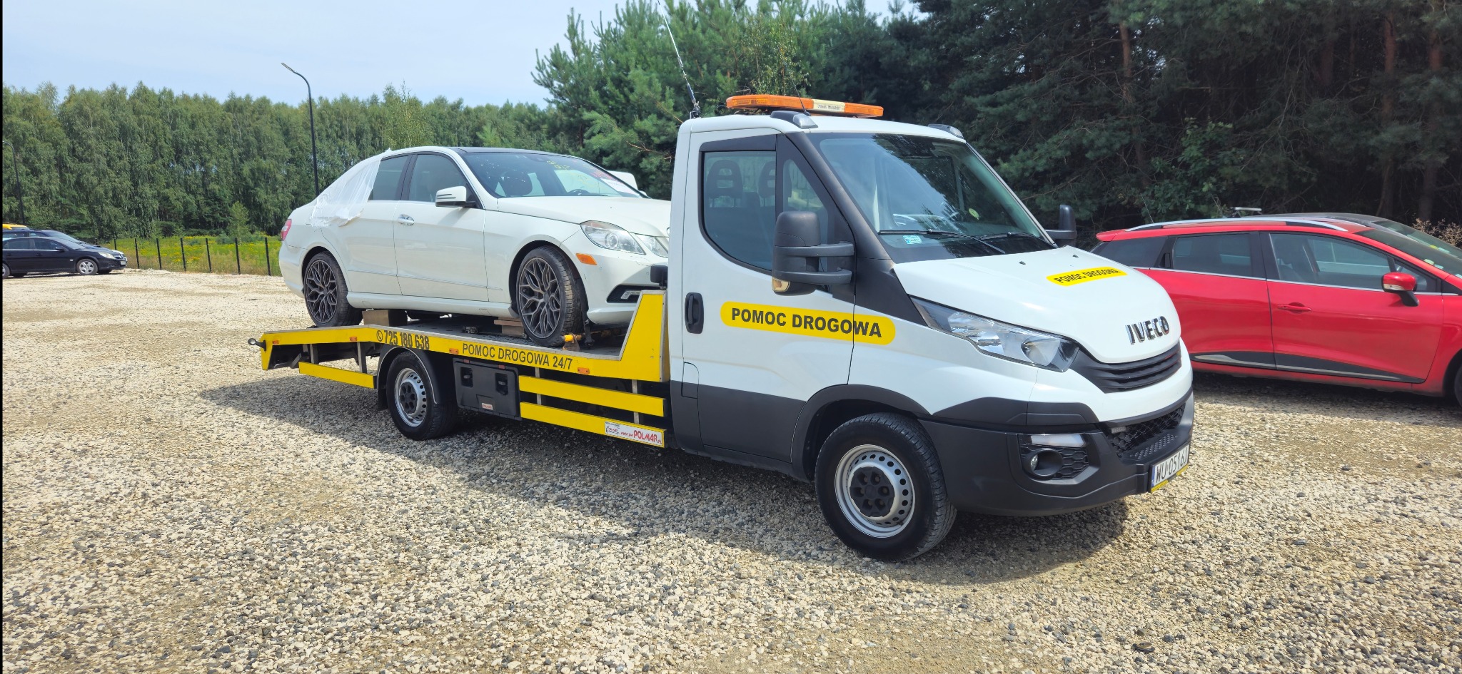 Biała pomoc drogowa Iveco z pomarańczowym światłem ostrzegawczym przewozi biały samochód osobowy z folią ochronną na szybach, na żwirowym parkingu z drzewami w tle.