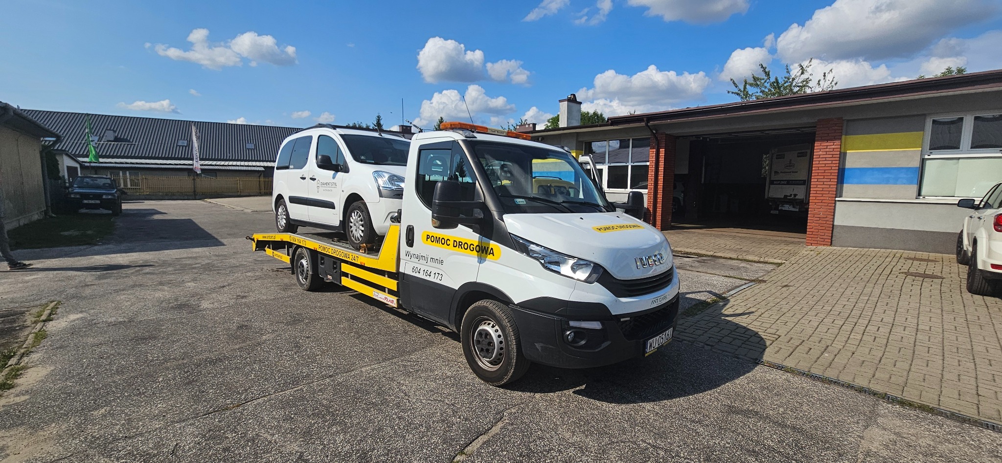 Biała ciężarówka pomocy drogowej z pomarańczowym światłem ostrzegawczym przewozi biały samochód dostawczy marki Citroen na żółtej platformie, na tle budynków i błękitnego nieba z chmurami.