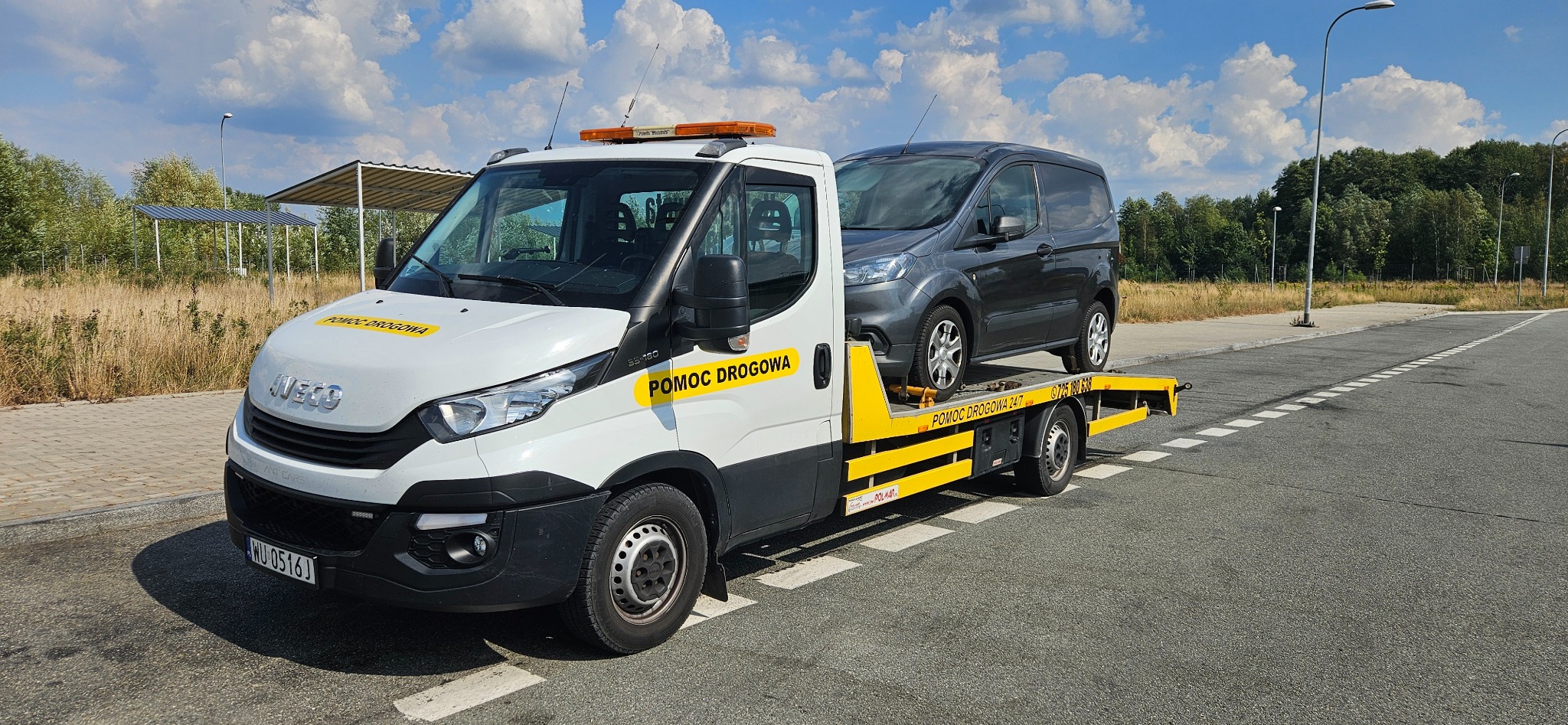 Biała ciężarówka pomocy drogowej marki Iveco z żółtymi elementami i napisem 'POMOC DROGOWA' transportuje szarego vana na platformie. Widoczne światła ostrzegawcze na dachu pojazdu pomocy drogowej.