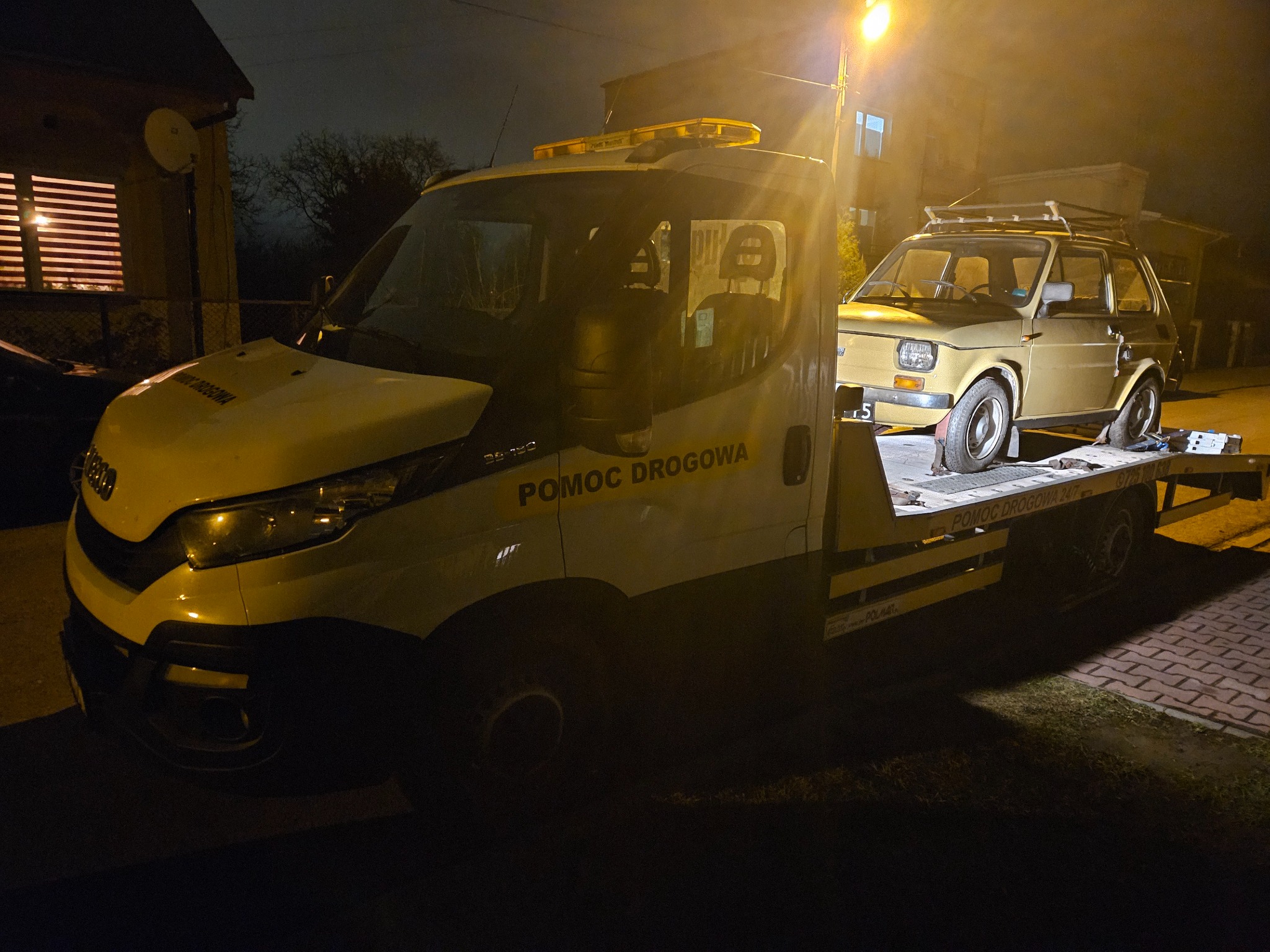 Żółty Fiat 126p z bagażnikiem dachowym transportowany na platformie pojazdu pomocy drogowej Iveco w nocnych warunkach oświetleniowych.