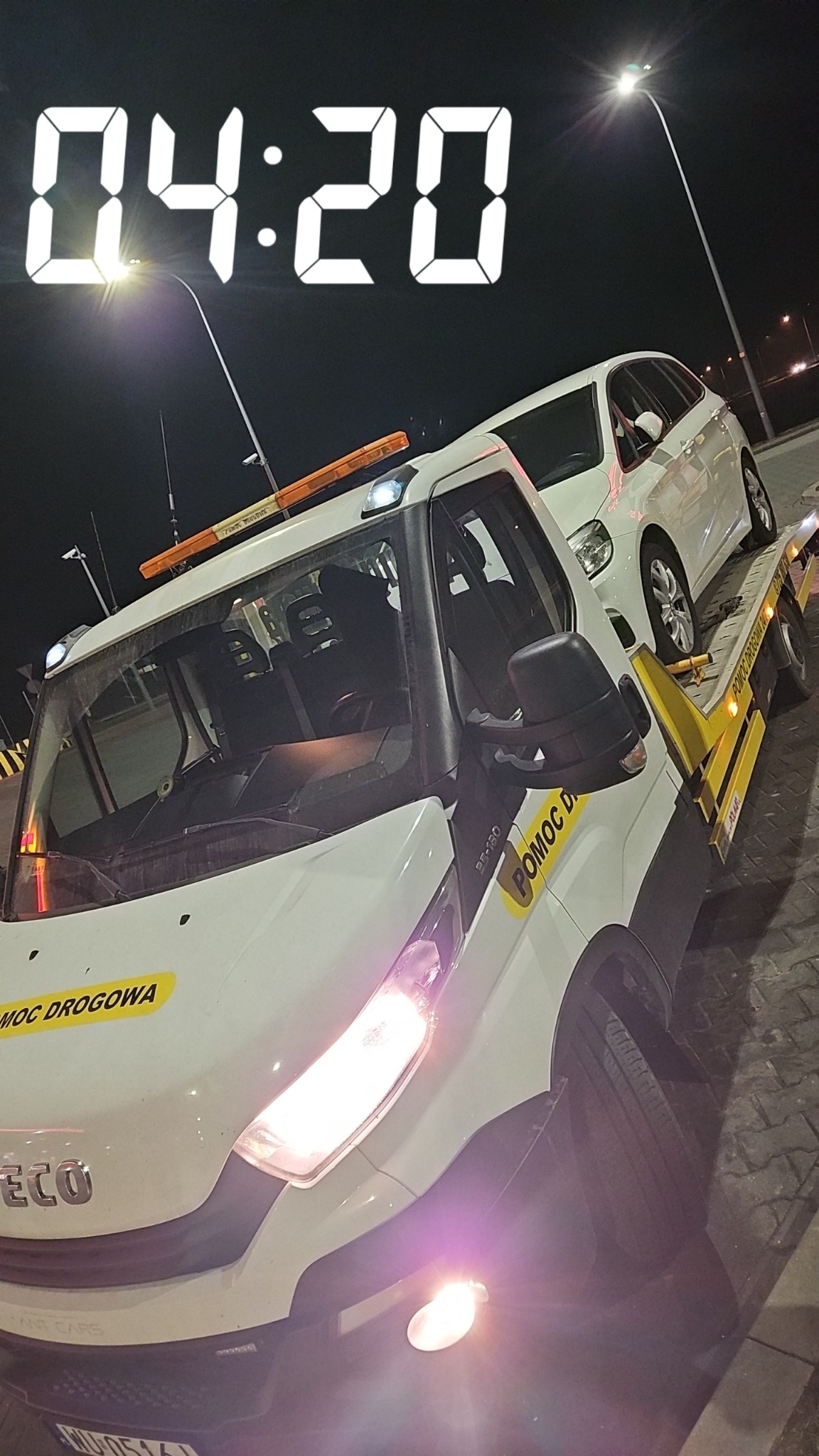 Biała laweta z przewożonym białym samochodem osobowym na tle nocnego krajobrazu w Siedliszczach, widoczne światła uliczne i cyfrowy zegar 04:20 nałożony na zdjęcie.