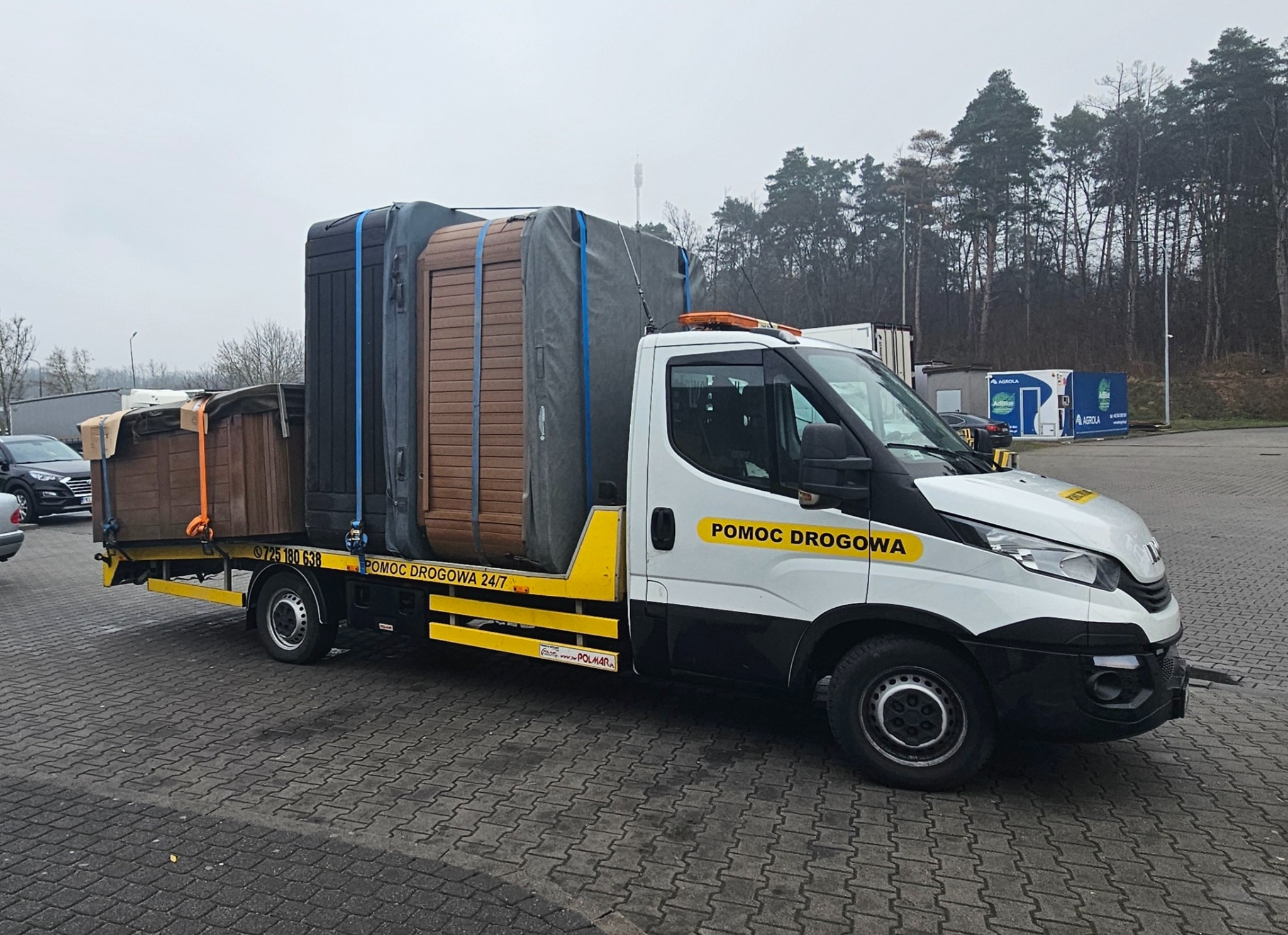 Pomoc drogowa przewozi na platformie kilka dużych, zabezpieczonych plandekami i pasami wanien spa w różnych kolorach, widoczne numery kontaktowe na pojeździe.