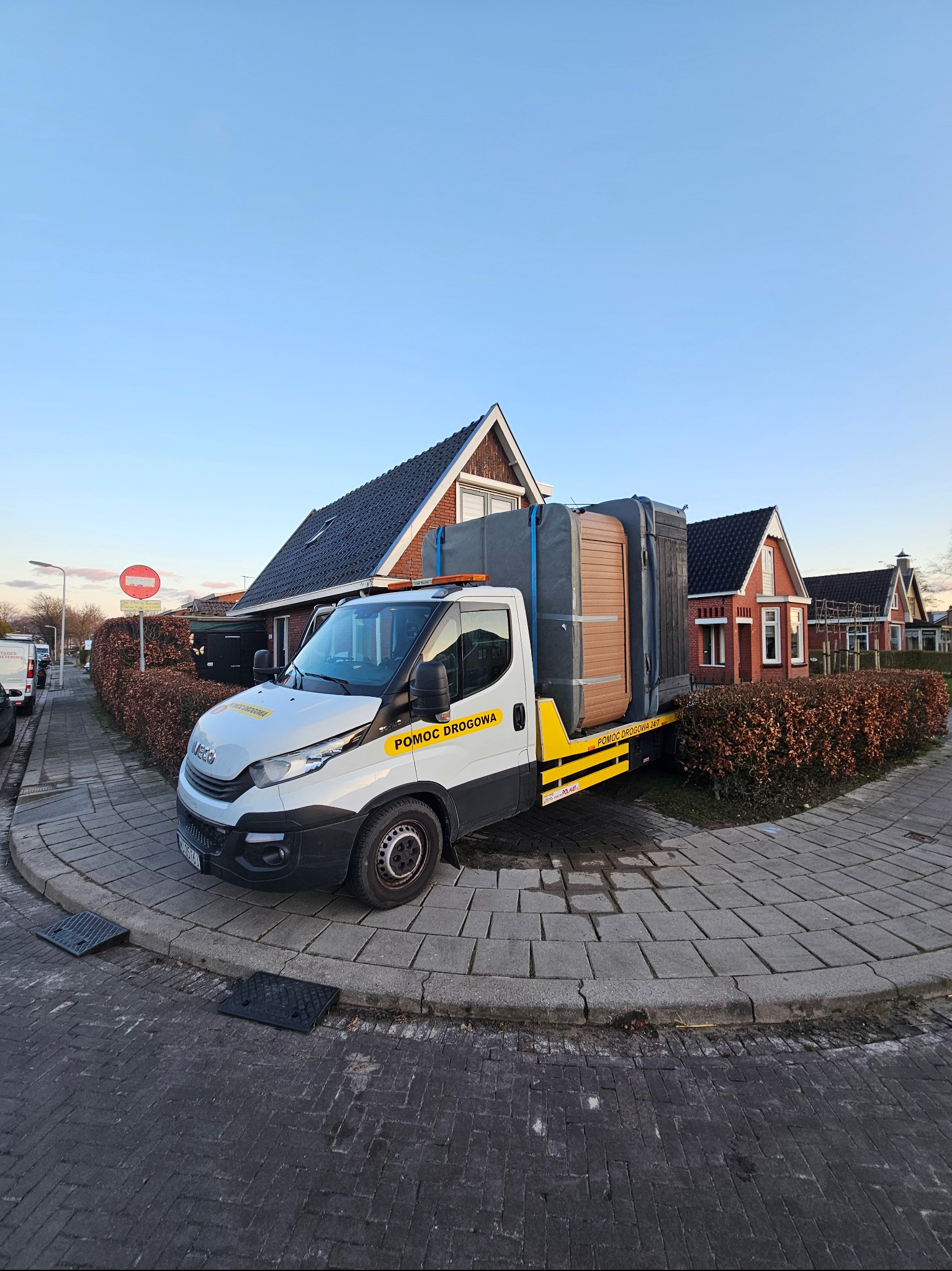 Biała ciężarówka pomocy drogowej z żółtymi elementami transportuje zabezpieczone folią jacuzzi na tle domów jednorodzinnych w spokojnej okolicy.