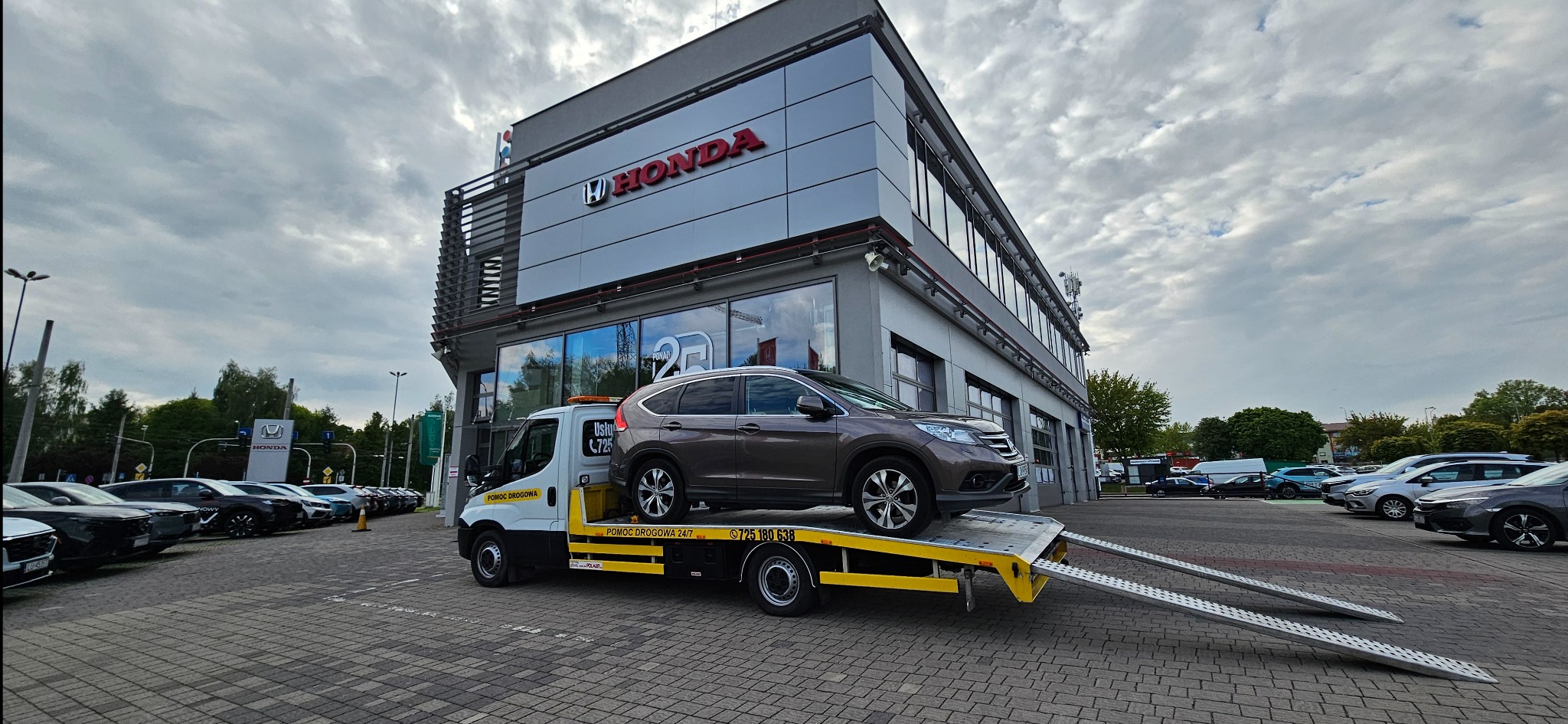 Brązowa Honda SUV na żółtej lawecie przed salonem Hondy. Pomoc drogowa 24h, usługi transportowe. Samochody na parkingu w tle, pochmurne niebo.