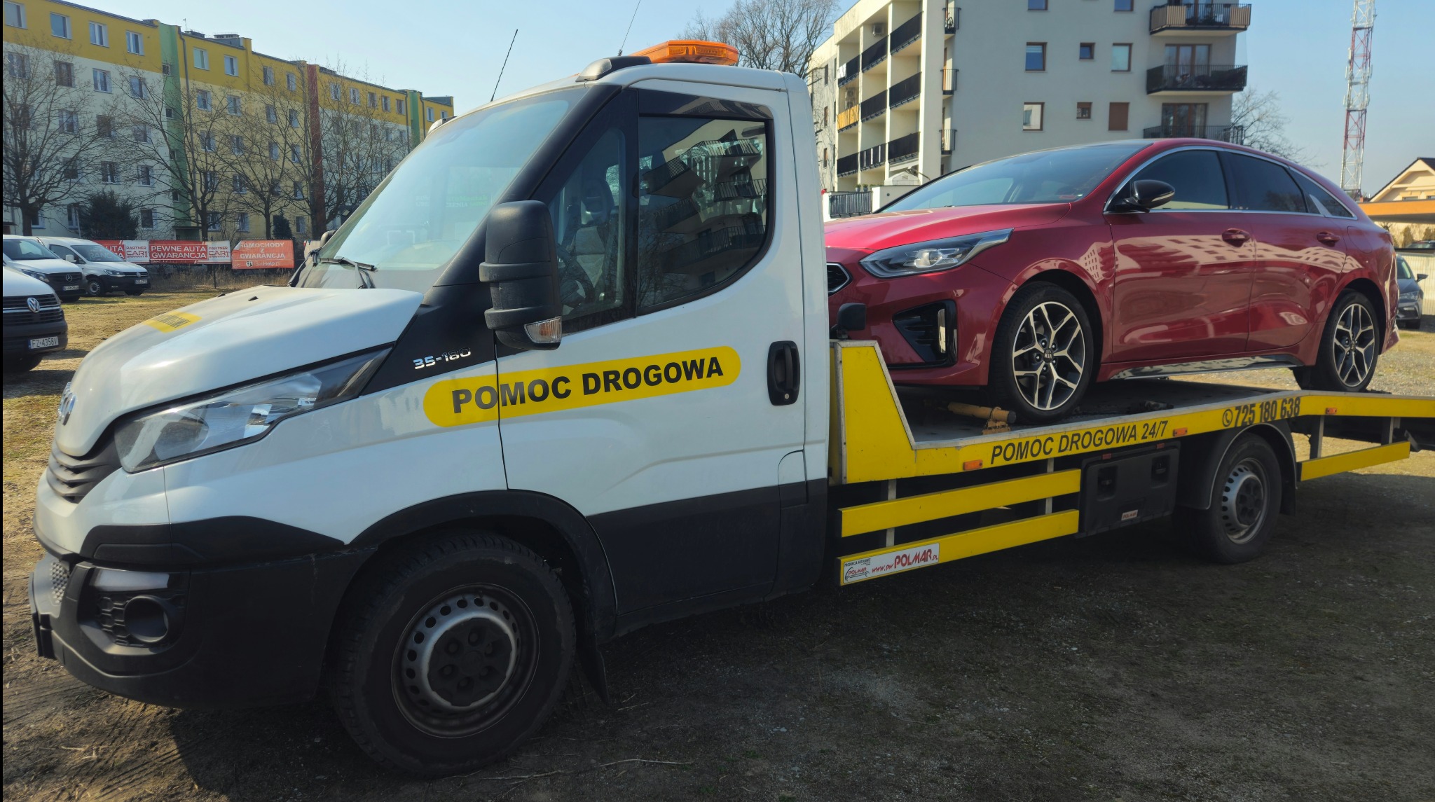 Laweta z czerwonym autem na platformie, w Siedliszczach. Żółte oznaczenia 'Pomoc Drogowa' na białej kabinie. W tle budynki mieszkalne i błękitne niebo.