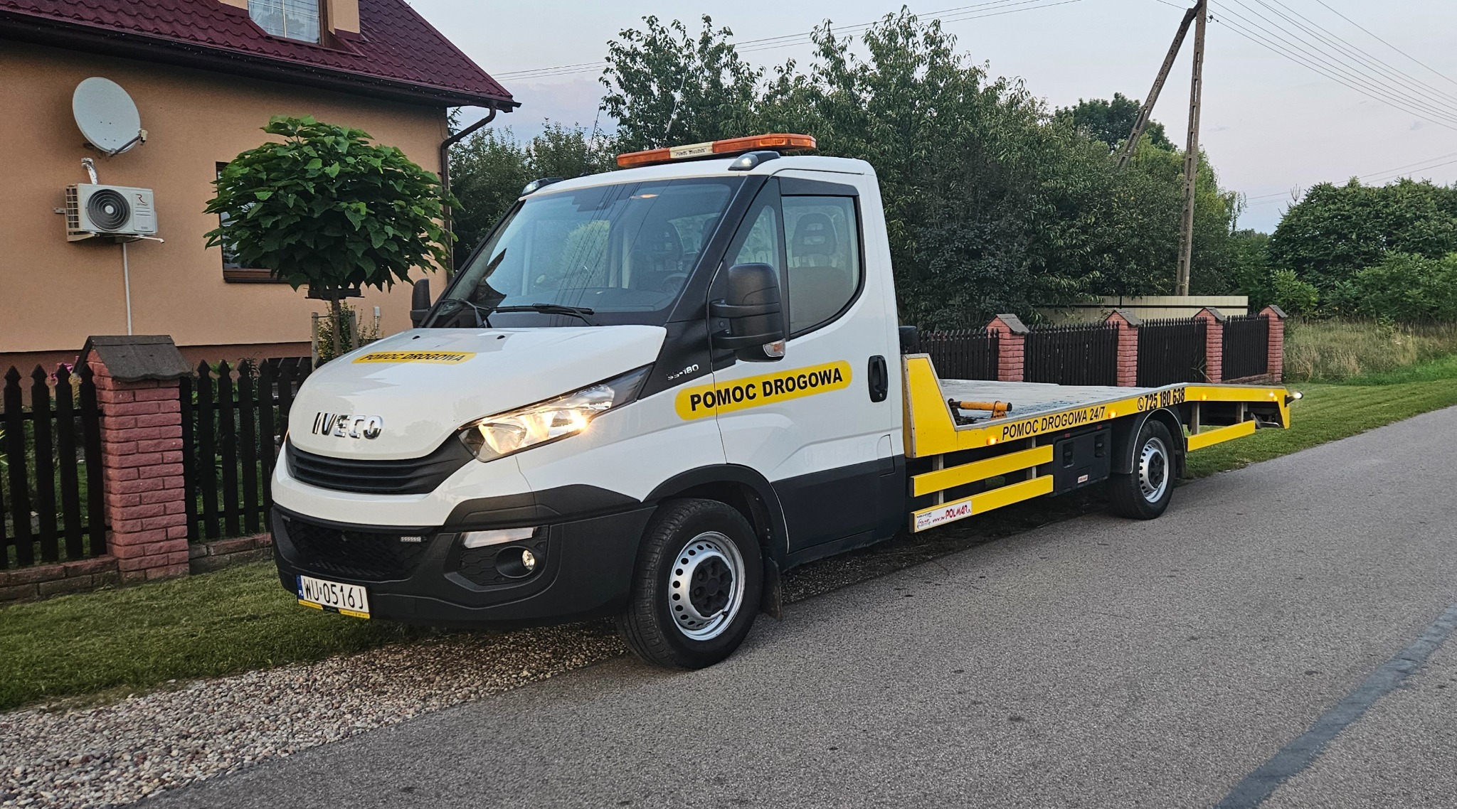 Biała laweta Iveco z żółtymi elementami i napisem 'Pomoc Drogowa' zaparkowana na poboczu drogi, na tle domu i zieleni. Widoczny numer telefonu.