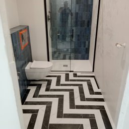 Robert M-k - Wykończona łazienka z kabiną prysznicową typu walk-in, podwieszaną toaletą i podłogą w czarno-biały wzór jodełki; na ścianach białe i niebieskie płytki.