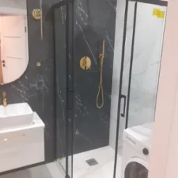 Nowoczesna łazienka z kabiną prysznicową w czarnej ramie, złotą armaturą na tle czarnego marmuru, białą umywalką nablatową i pralką.