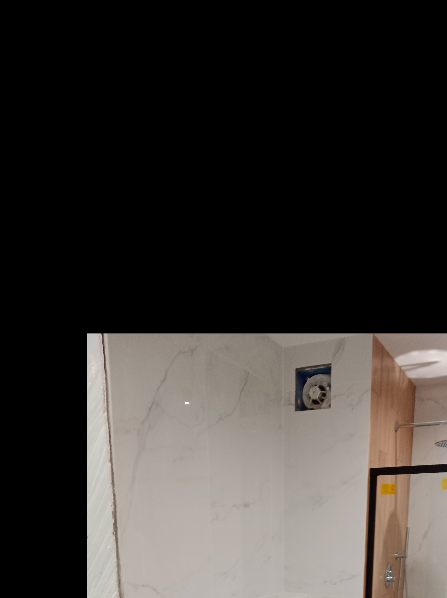 Wykończona łazienka z białymi płytkami imitującymi marmur, drewnianą ścianką działową, szklaną kabiną prysznicową i wentylatorem w ścianie.