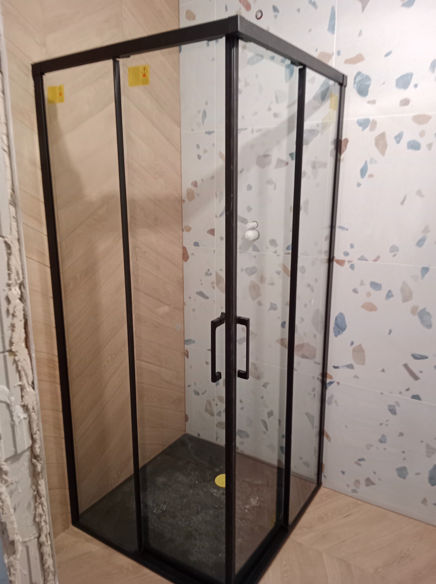 Nowoczesna kabina prysznicowa z czarnymi profilami na tle płytek imitujących drewno i mozaiki terrazzo.