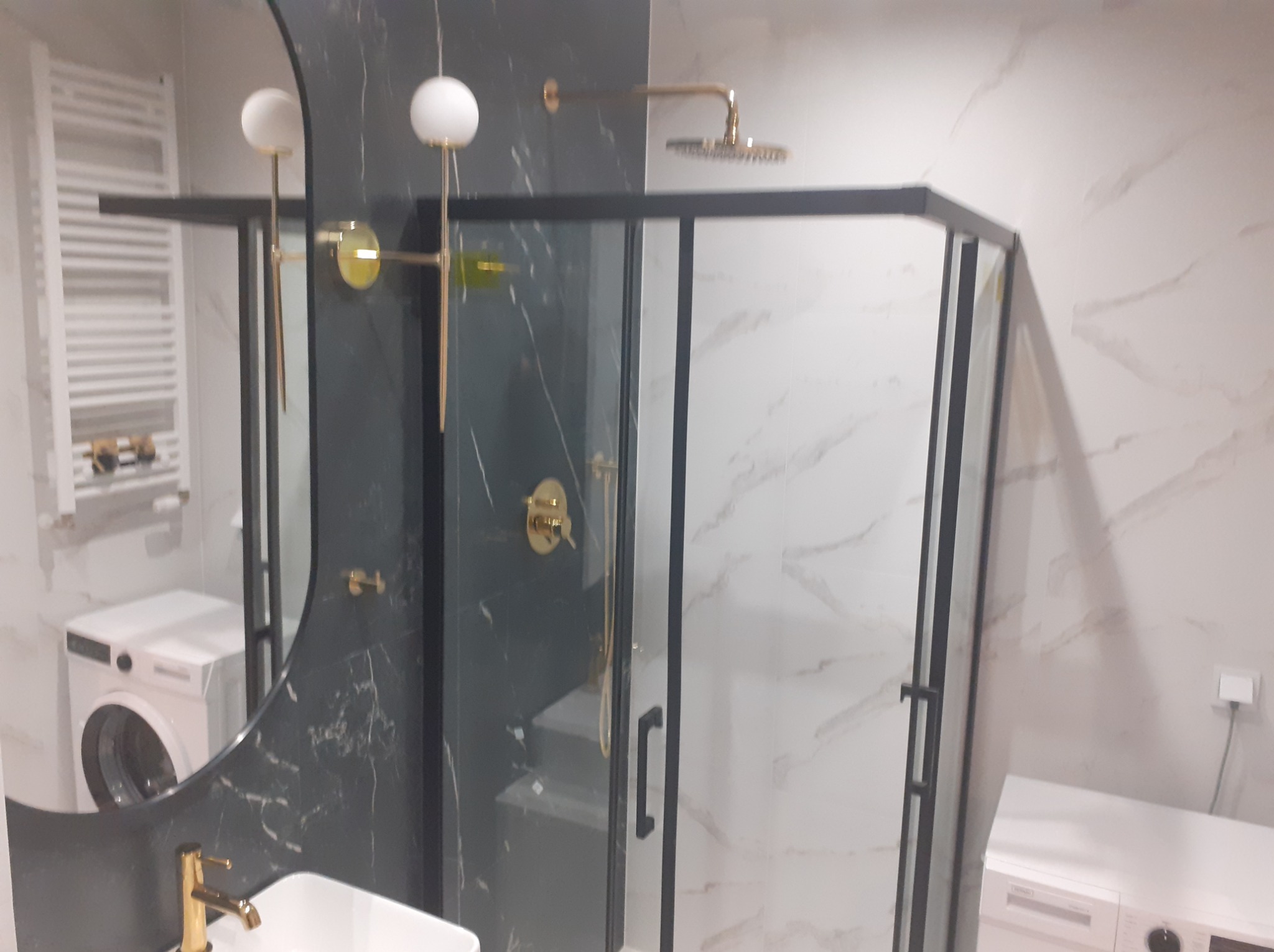 Nowoczesna łazienka z kabiną prysznicową w czarnej ramie, złotą armaturą i umywalką, widoczna pralka w tle.