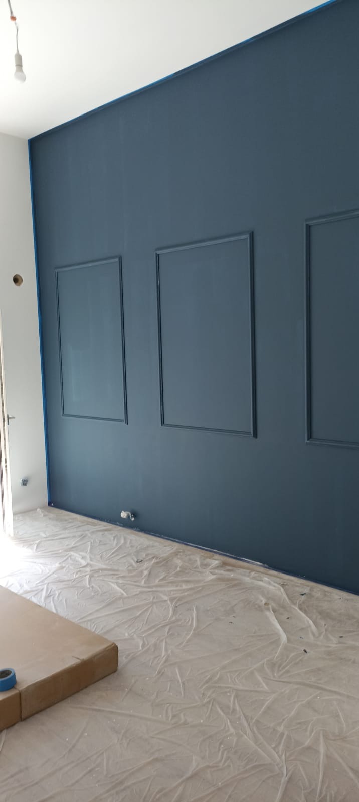 Ściana pomalowana na ciemny odcień niebieskiego z trzema dekoracyjnymi ramkami, podłoga zabezpieczona folią malarską, widoczne taśmy malarskie przy krawędzi ściany.