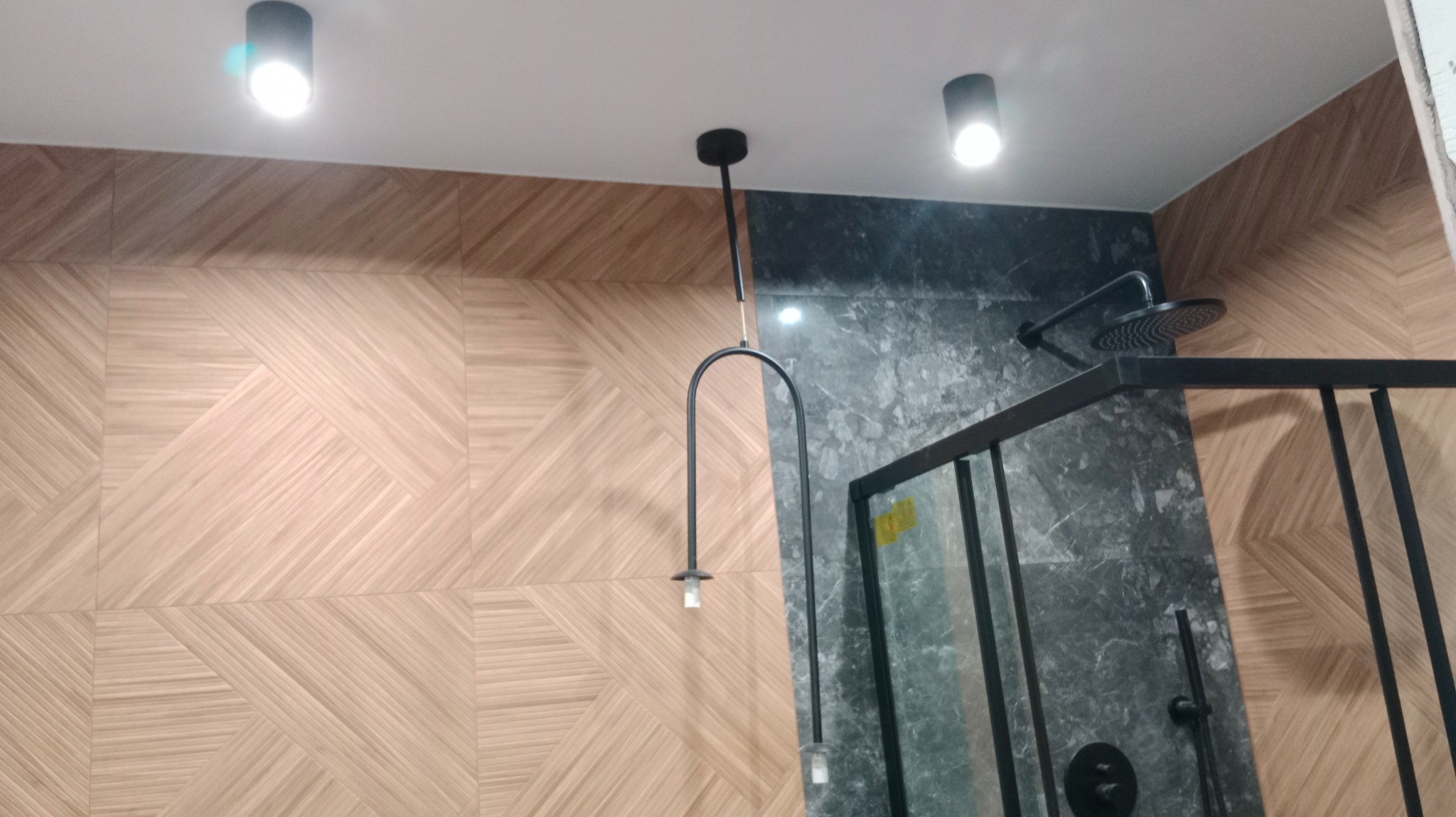 Nowoczesna łazienka z kabiną prysznicową typu walk-in, wykończona płytkami imitującymi drewno na jednej ścianie i szarym marmurem na drugiej, oświetlona trzema punktowymi lampami sufitowymi...