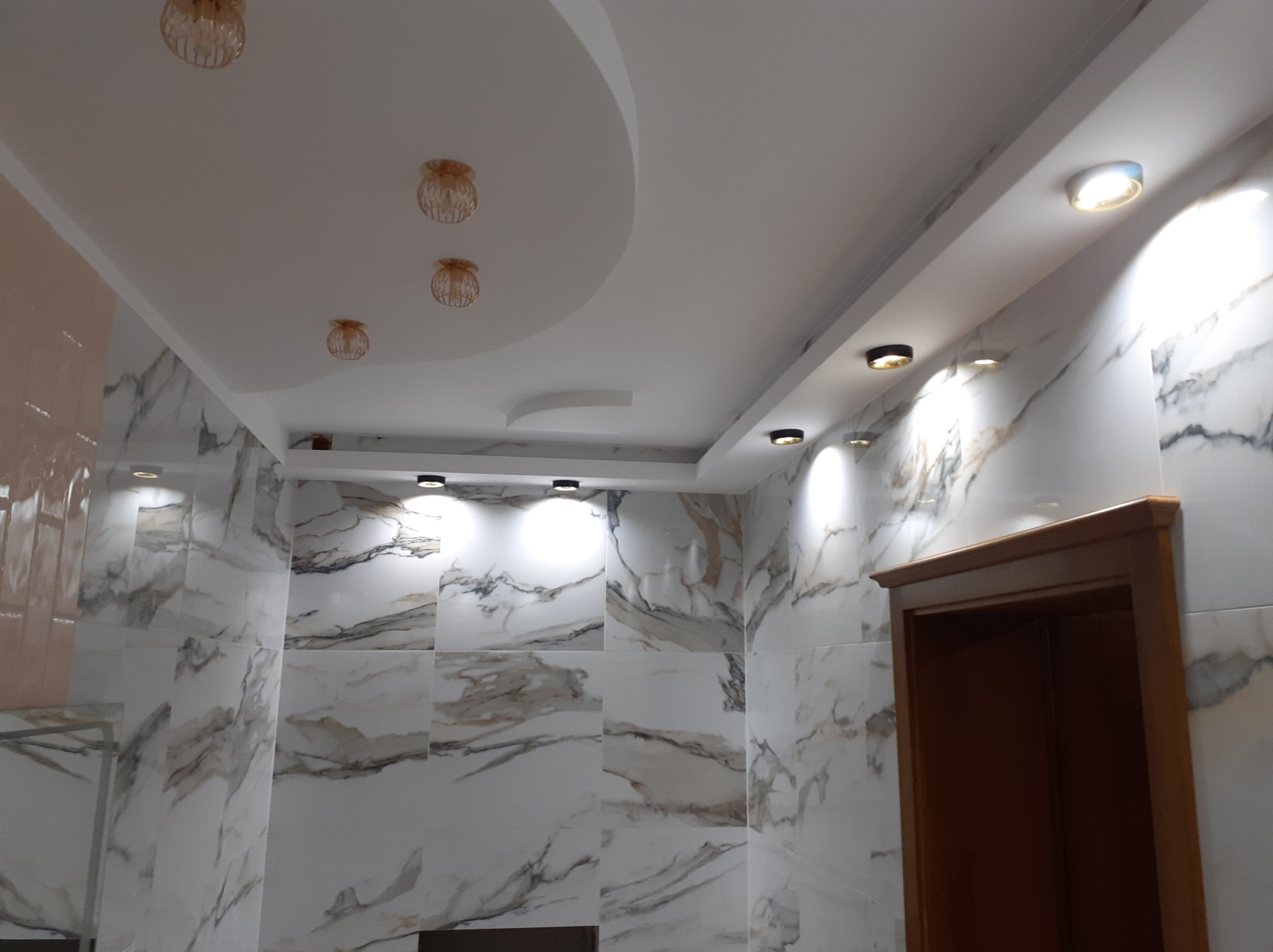 Nowoczesna łazienka z białymi płytkami imitującymi marmur, ozdobnym podwieszanym sufitem z oświetleniem punktowym i dekoracyjnymi lampami wiszącymi.