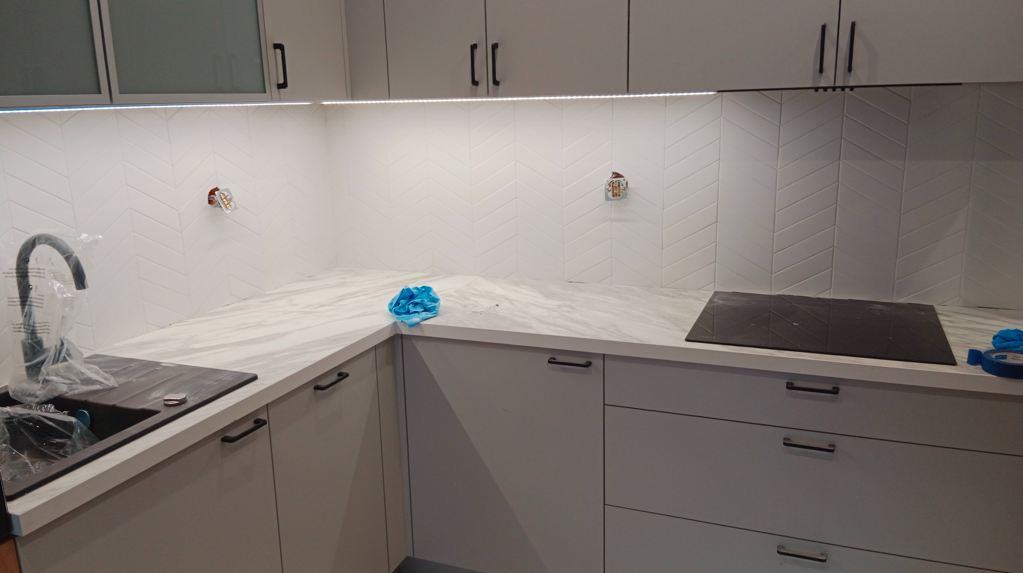 Nowoczesna kuchnia w trakcie montażu: białe płytki ułożone w jodełkę na ścianie, marmurowy blat, szare szafki z czarnymi uchwytami, widoczne puszki elektryczne bez osprzętu, umywalka zakryta folią...