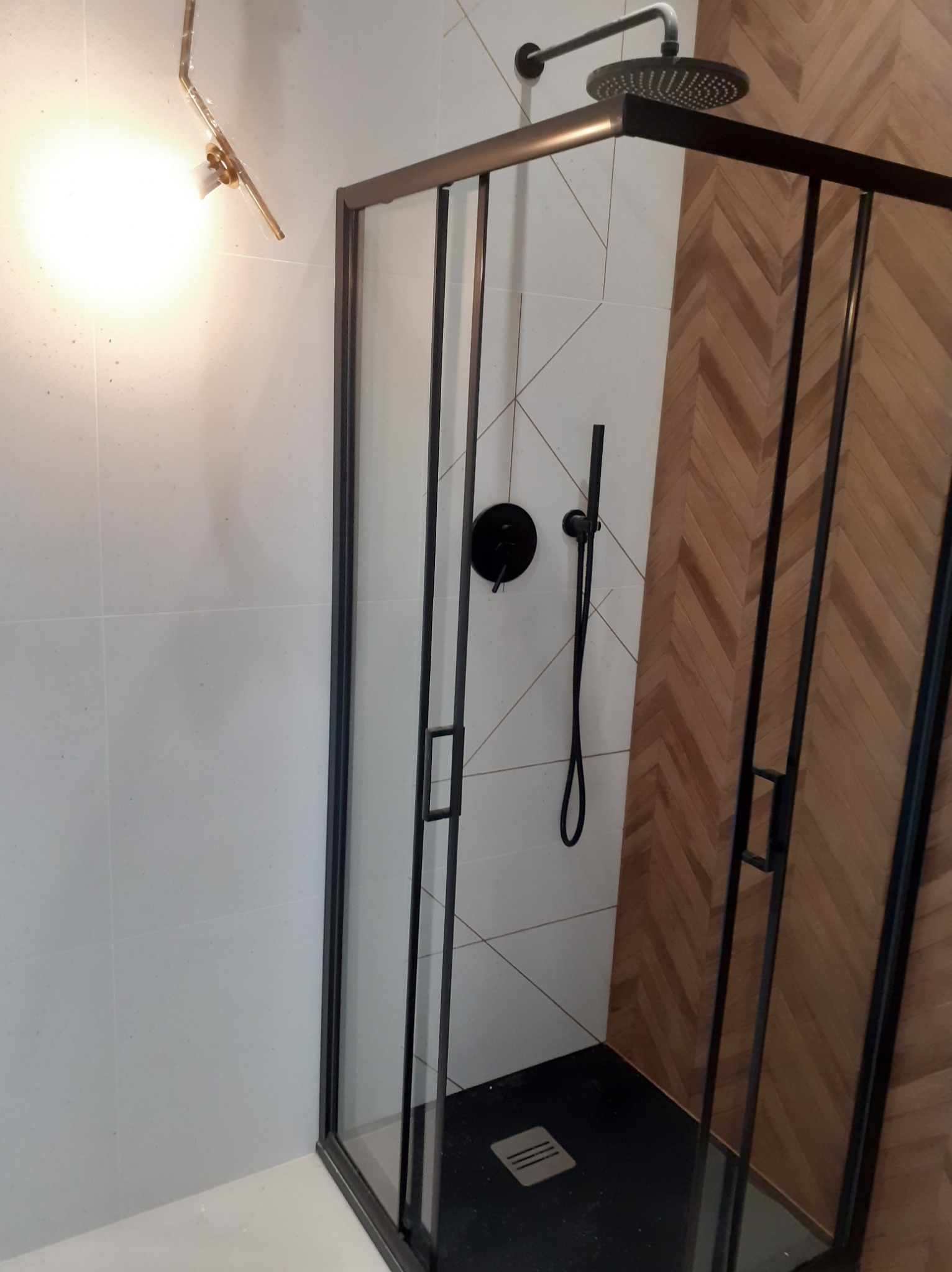 Nowoczesna kabina prysznicowa z czarnymi profilami, deszczownicą i armaturą na tle ściany wyłożonej białymi płytkami i drewnopodobną mozaiką ułożoną w jodełkę.