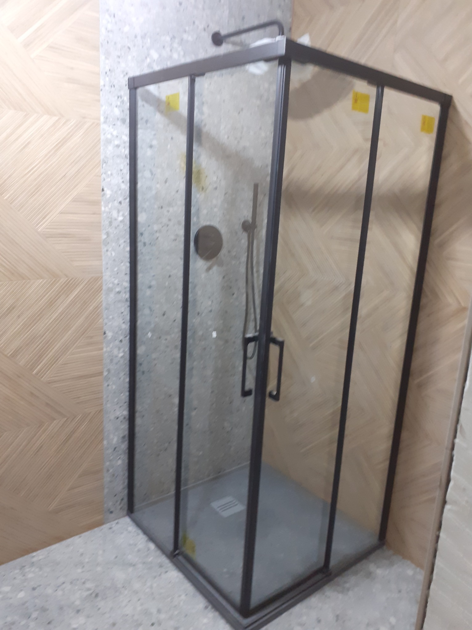 Szklana kabina prysznicowa z czarnymi profilami, zamontowana w łazience wykończonej płytkami imitującymi drewno i lastryko.