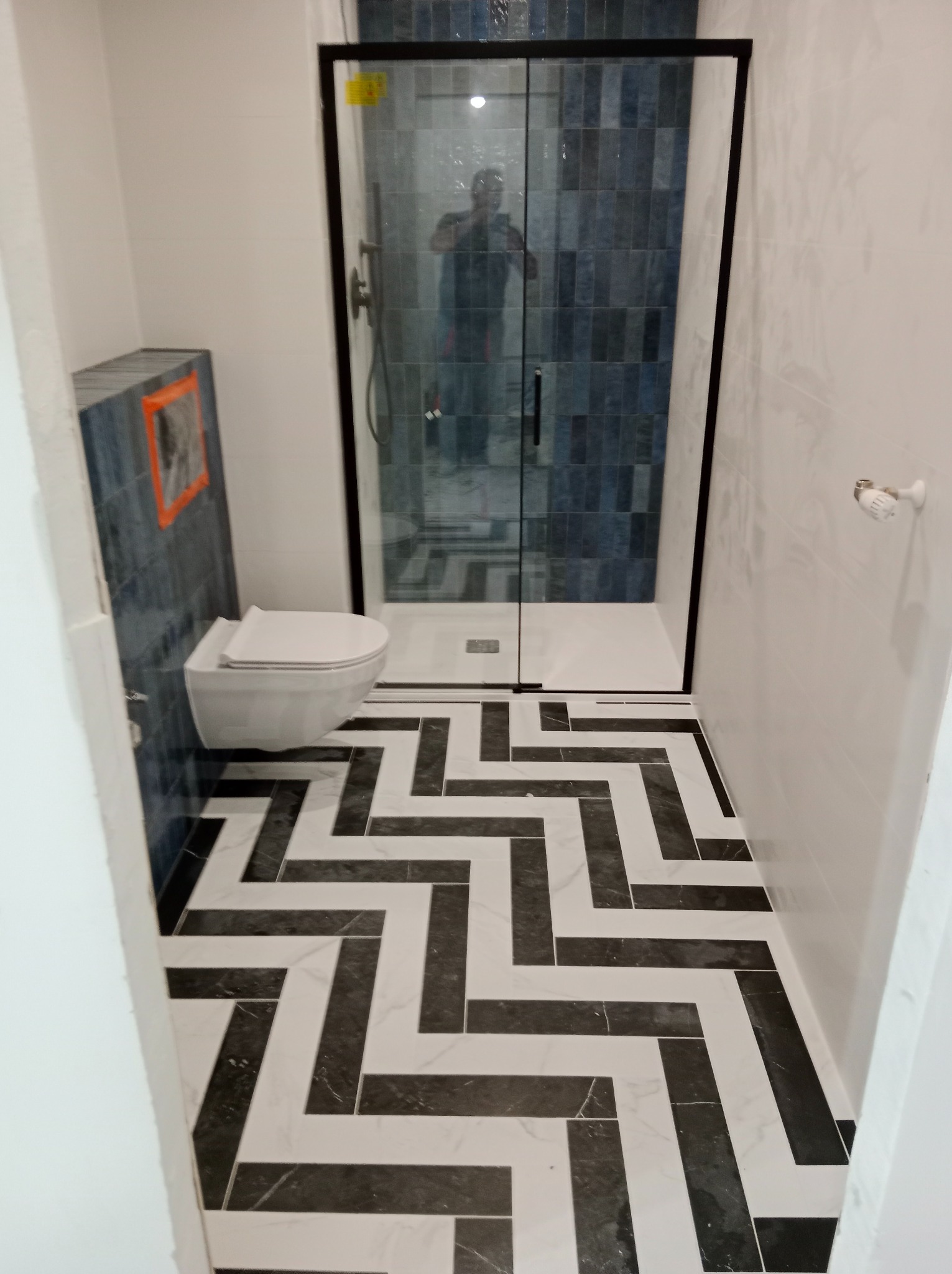 Wykończona łazienka z kabiną prysznicową typu walk-in, podwieszaną toaletą i podłogą w czarno-biały wzór jodełki; na ścianach białe i niebieskie płytki.