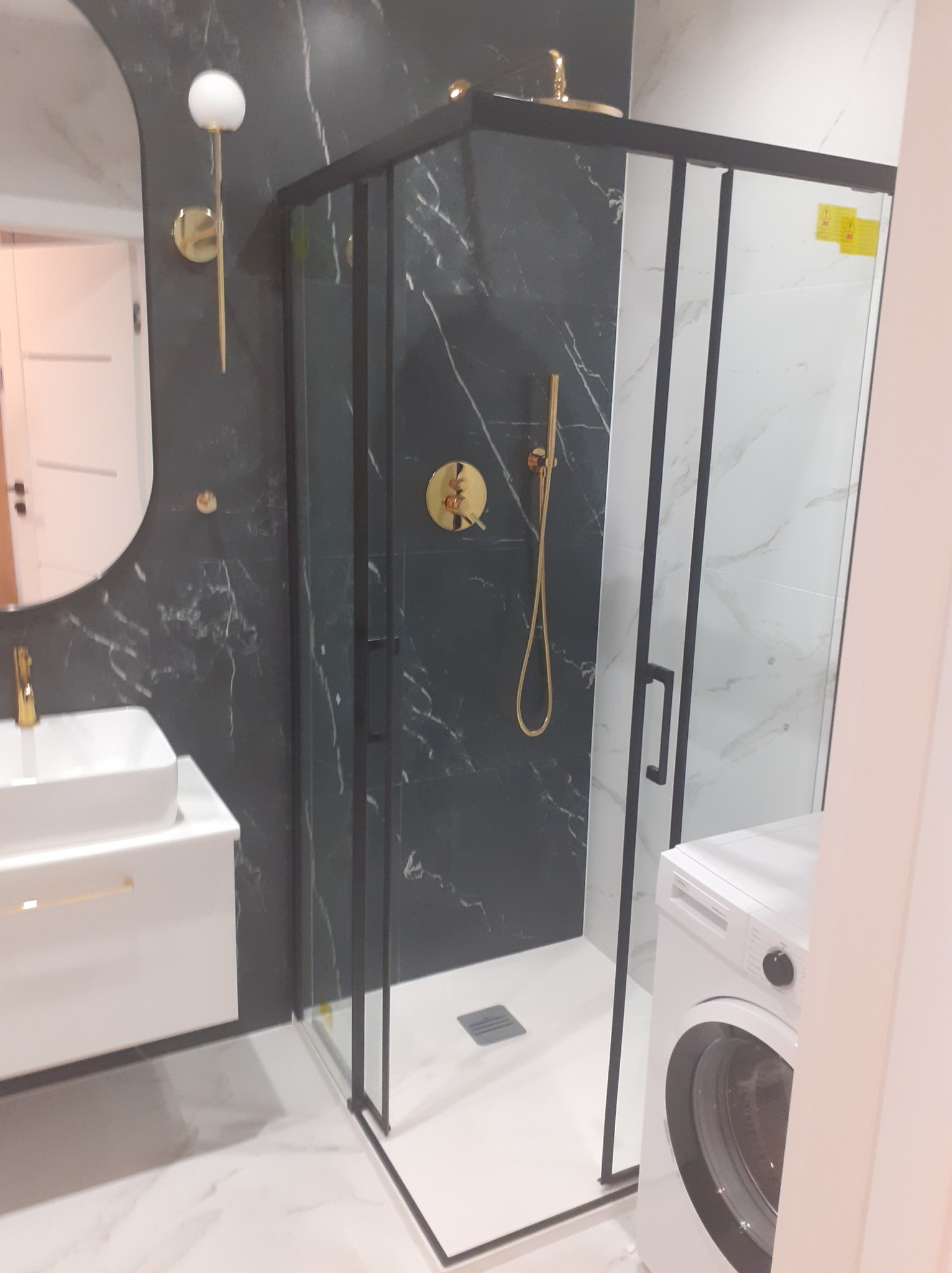 Nowoczesna łazienka z kabiną prysznicową w czarnej ramie, złotą armaturą na tle czarnego marmuru, białą umywalką nablatową i pralką.