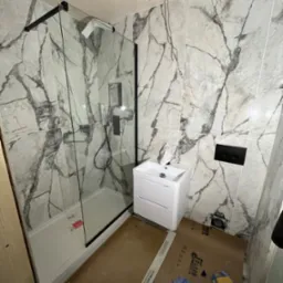 Łazienka w trakcie remontu: kabina prysznicowa z czarnymi ramami i marmurkowymi płytkami, biała umywalka z szafką, widoczne narzędzia i materiały na podłodze, niedokończone podłączenie elektryczne.