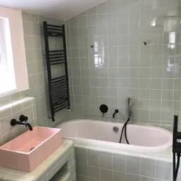 Wykończona łazienka w pastelowych odcieniach zieleni, z wanną obudowaną kafelkami, różową umywalką na postumencie z kafelków, czarną armaturą i grzejnikiem drabinkowym.