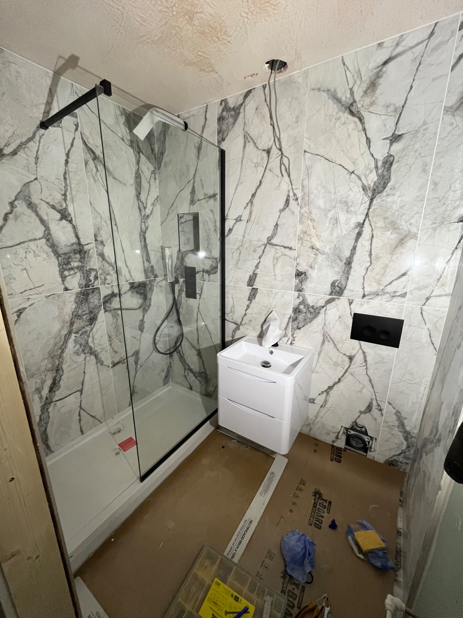 Łazienka w trakcie remontu: kabina prysznicowa z czarnymi ramami i marmurkowymi płytkami, biała umywalka z szafką, widoczne narzędzia i materiały na podłodze, niedokończone podłączenie elektryczne.