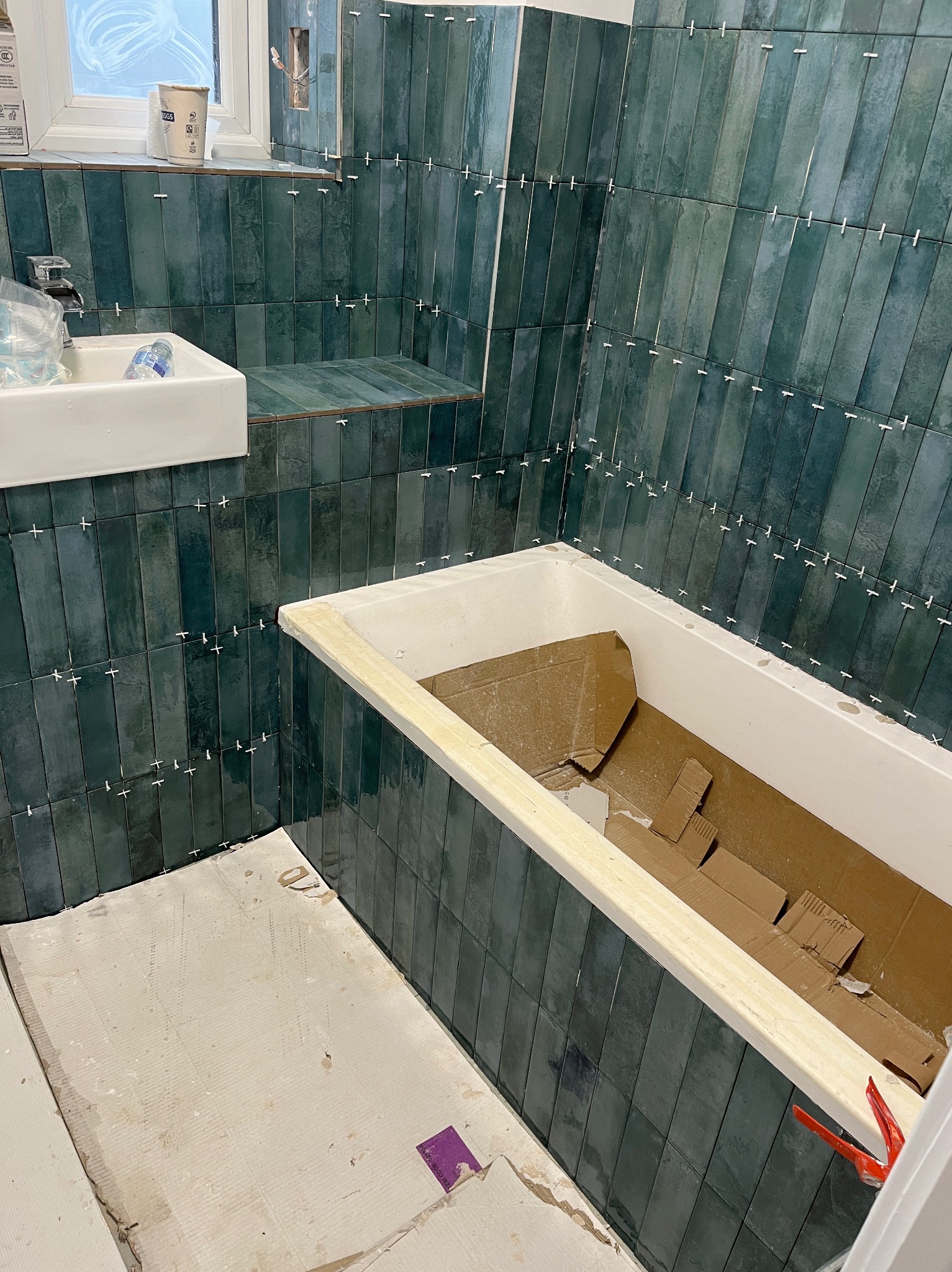 Łazienka w trakcie remontu: pionowo ułożone, ciemnozielone płytki na ścianach z białymi krzyżykami dystansowymi, wanna zabezpieczona kartonem, umywalka i surowa podłoga.
