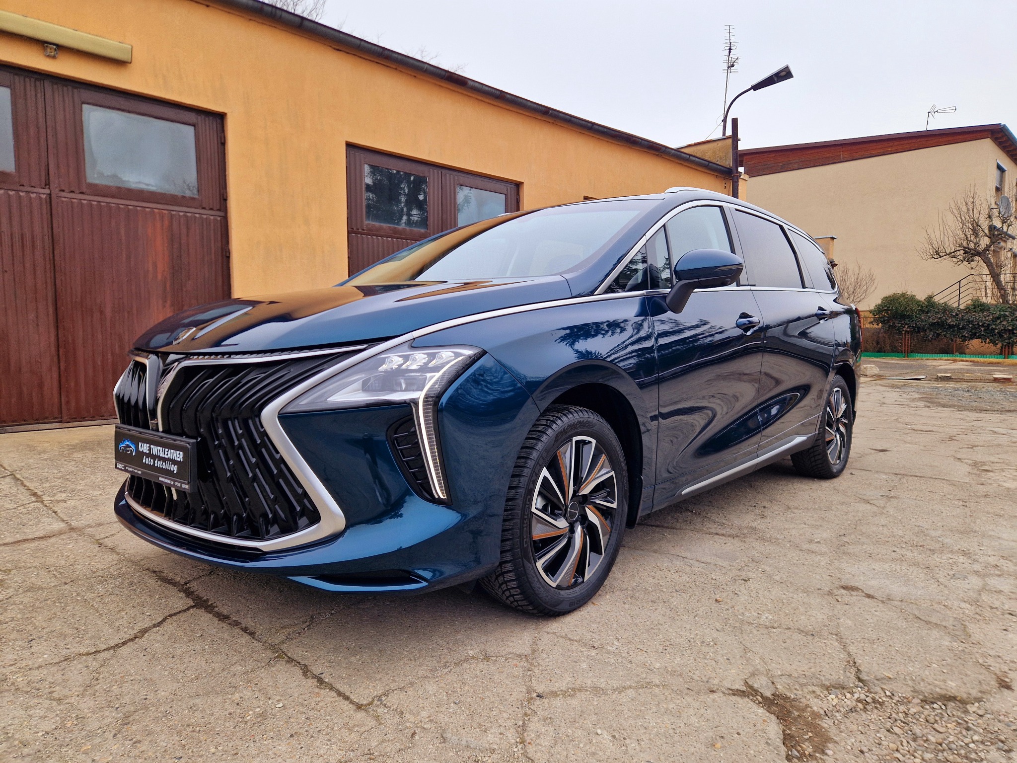 Błyszczący, granatowy minivan po detailingu, zaparkowany przed budynkiem z brązowymi wrotami garażowymi; widoczny logo firmy detailingowej na tabliczce.