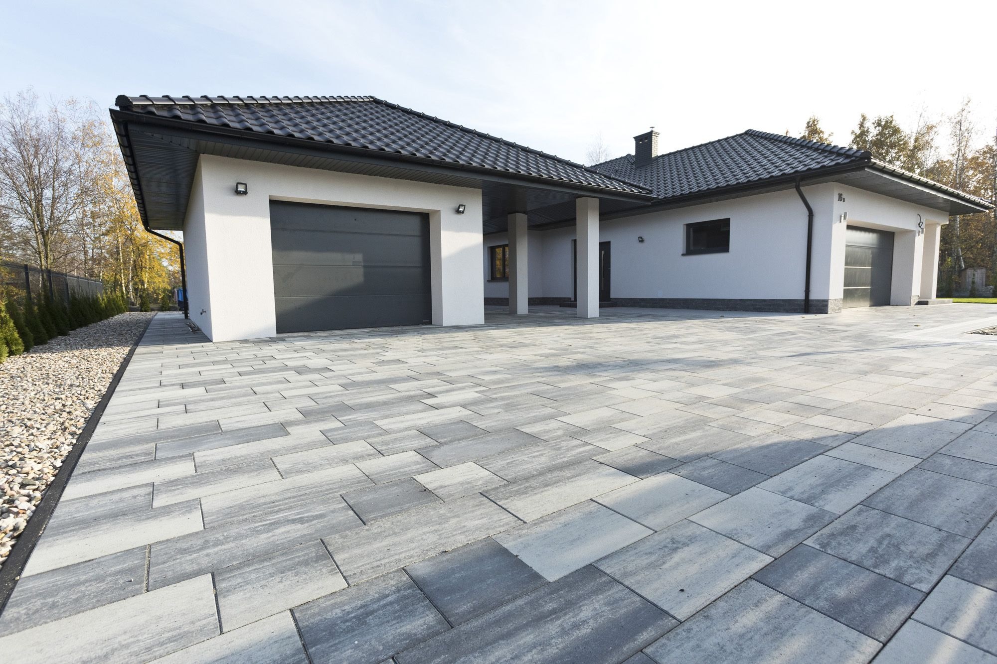 Szeroki podjazd do nowoczesnego domu z dwoma garażami, wyłożony szarą kostką w prostokątnym wzorze, z widocznymi elementami wykończeniowymi i zagospodarowaniem terenu.