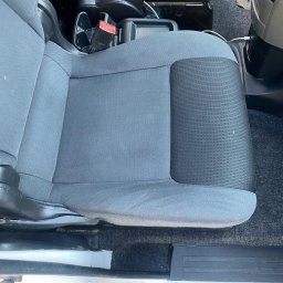 BÓBR CLEANING OLHA BEBRISA - Szare, materiałowe siedzenie samochodowe z widocznymi plamami, zapięciem pasów i elementami konsoli środkowej.