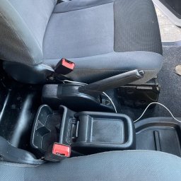 BÓBR CLEANING OLHA BEBRISA - Szare, materiałowe siedzenie samochodowe z widocznym zapięciem pasa, czarnym podłokietnikiem i fragmentem podłogi. Widoczny kabel pod fotelem.