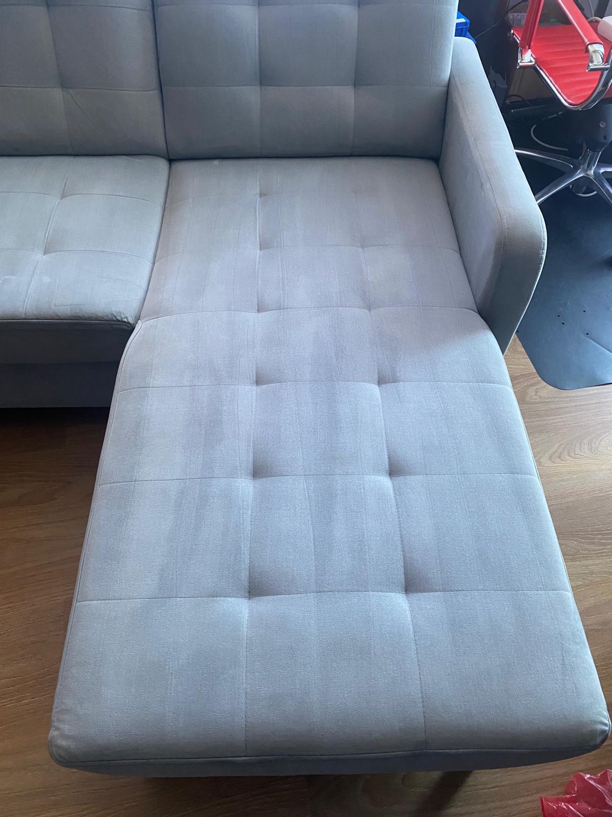 Jasnoszara, narożna kanapa z wyraźnym podziałem na sekcje, widoczne ślady po czyszczeniu, częściowo odświeżony wygląd, podłoga z paneli w tle.