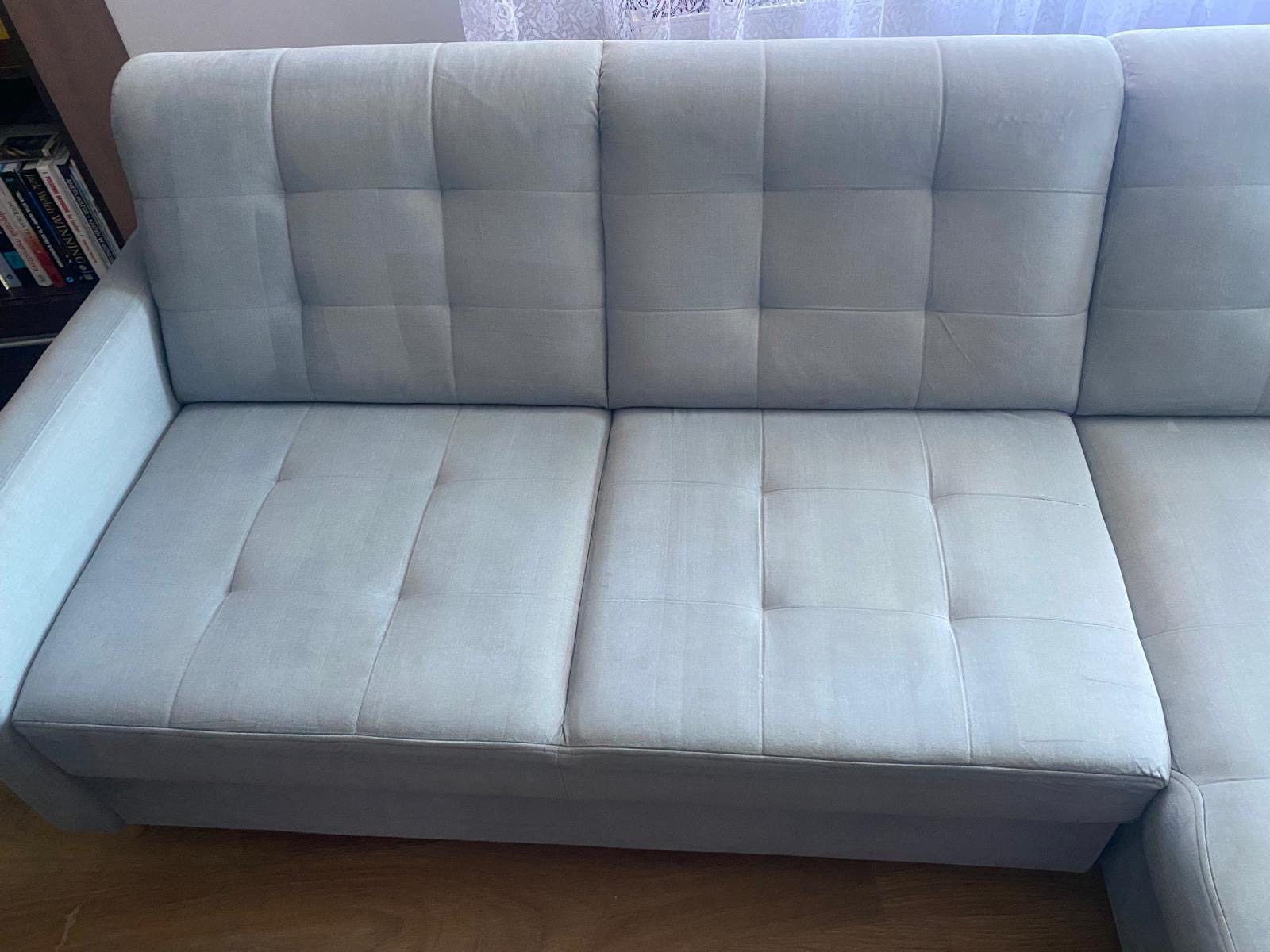 Jasnoszara, rozłożona sofa z tapicerowanymi kwadratami, widoczna z bliska, na drewnianej podłodze, z półką na książki w tle.