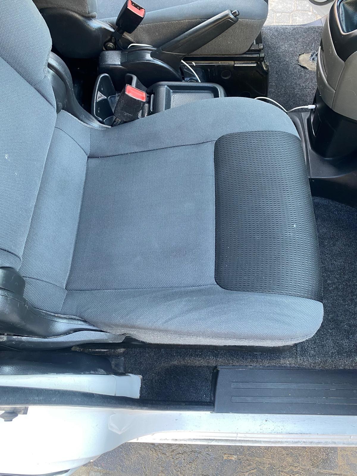 Szare, materiałowe siedzenie samochodowe z widocznymi plamami, zapięciem pasów i elementami konsoli środkowej.