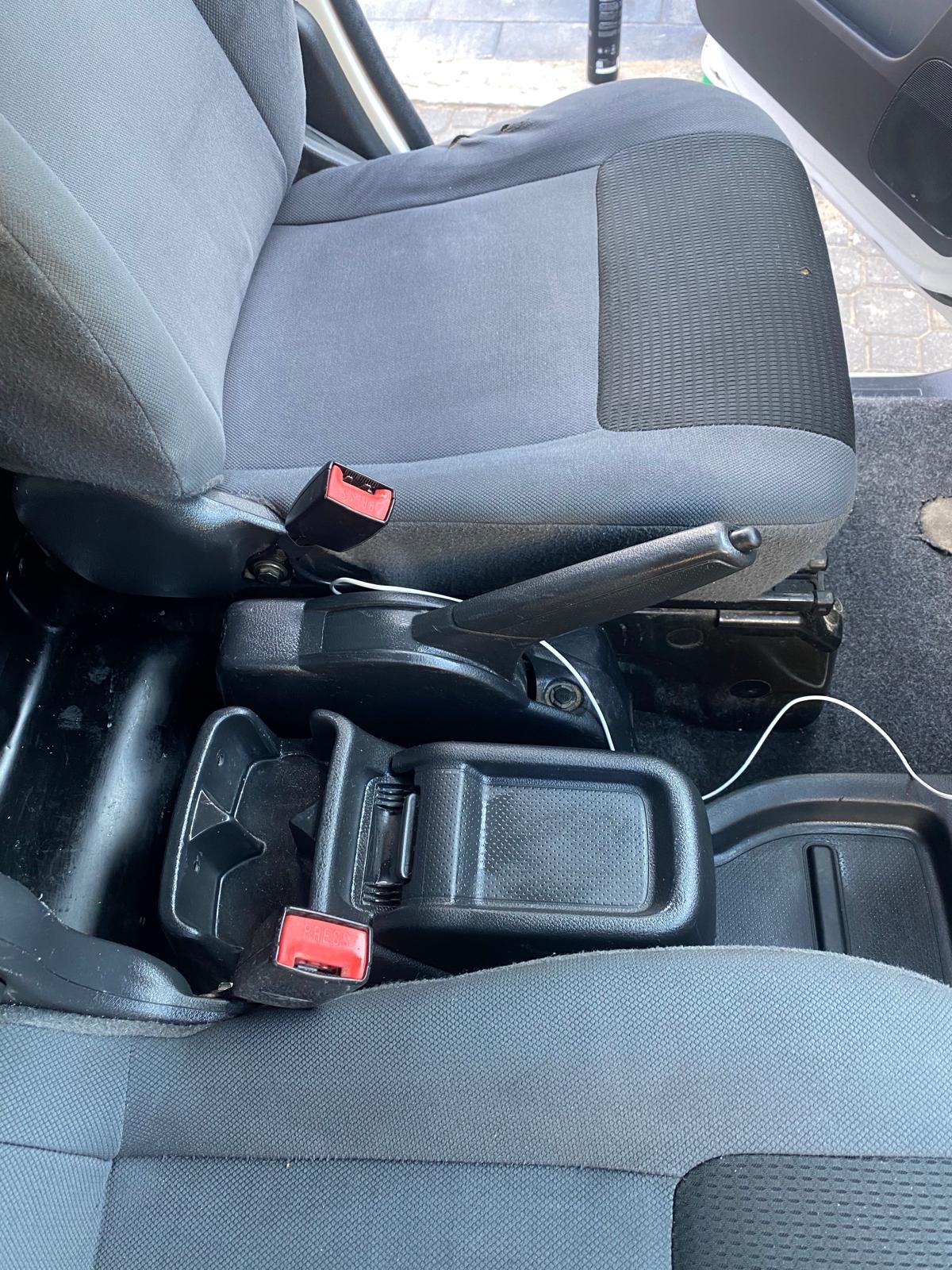 Szare, materiałowe siedzenie samochodowe z widocznym zapięciem pasa, czarnym podłokietnikiem i fragmentem podłogi. Widoczny kabel pod fotelem.