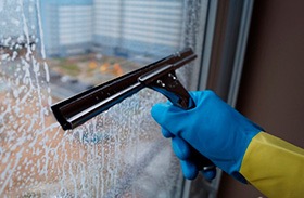 Ręka w niebieskiej rękawicy czyści szybę okienną za pomocą ściągaczki, pozostawiając smugi piany. Widok na rozmyte budynki za oknem.