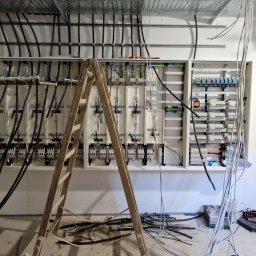 M&D-INSTAL SPÓŁKA Z OGRANICZONĄ ODPOWIEDZIALNOŚCIĄ - Szafa rozdzielcza z licznymi przewodami elektrycznymi w surowym wnętrzu, widoczna drewniana drabina i narzędzia.