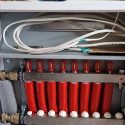 M&D-INSTAL SPÓŁKA Z OGRANICZONĄ ODPOWIEDZIALNOŚCIĄ - Rozdzielacz ogrzewania podłogowego z czerwonymi i niebieskimi rurami, widoczne okablowanie elektryczne w ścianie i narzędzia na półce powyżej.