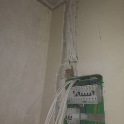 M&D-INSTAL SPÓŁKA Z OGRANICZONĄ ODPOWIEDZIALNOŚCIĄ - Ściana w surowym stanie z widocznymi przewodami elektrycznymi prowadzącymi do skrzynki rozdzielczej z zielonym obramowaniem, widoczne zabezpieczenia i kable wewnątrz skrzynki.