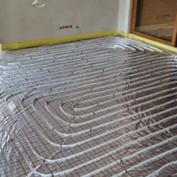 Białe rury ogrzewania podłogowego ułożone na czerwonej folii w surowym wnętrzu z szarymi ścianami i żółtą taśmą izolacyjną wzdłuż krawędzi.