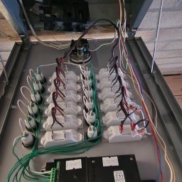 M&D-INSTAL SPÓŁKA Z OGRANICZONĄ ODPOWIEDZIALNOŚCIĄ - Otwarta skrzynka rozdzielcza z licznymi przewodami i złączkami, widoczne dwa czarne moduły RITO z zielonymi listwami zaciskowymi, dokumentacja Renz w tle.