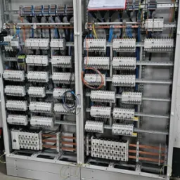 Dwie szafy elektryczne w trakcie montażu, widoczne liczne zabezpieczenia, przewody i listwy zaciskowe, na górze jednej z szaf dokumentacja techniczna.