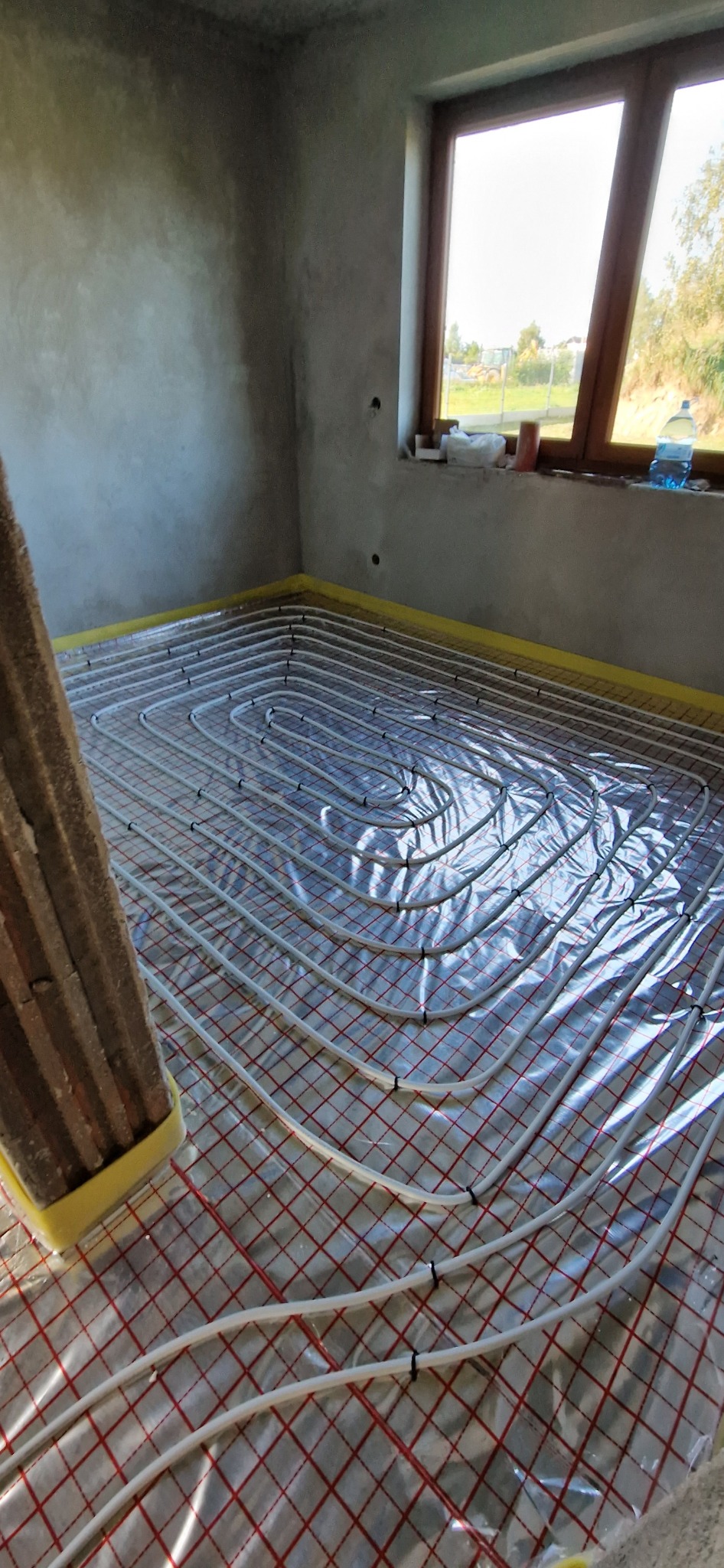 Montaż ogrzewania podłogowego: białe rurki ułożone w pętle na czerwonej siatce, przytwierdzone czarnymi klipsami, zabezpieczone folią, widok na surową podłogę i ścianę, okno w tle.