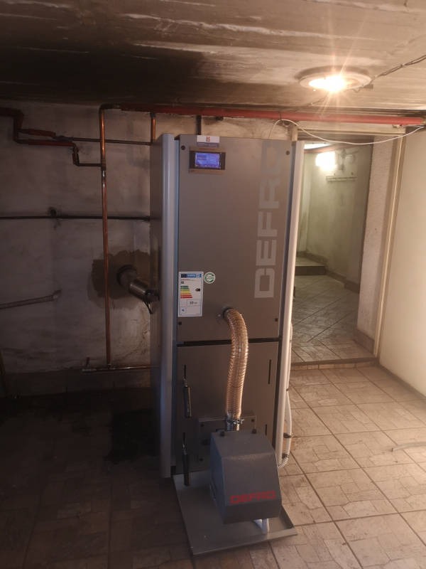 Nowoczesny piec na paliwo stałe marki Defro w piwnicy, widoczne rury instalacji grzewczej na ścianie, surowy betonowy strop z zaciekami.