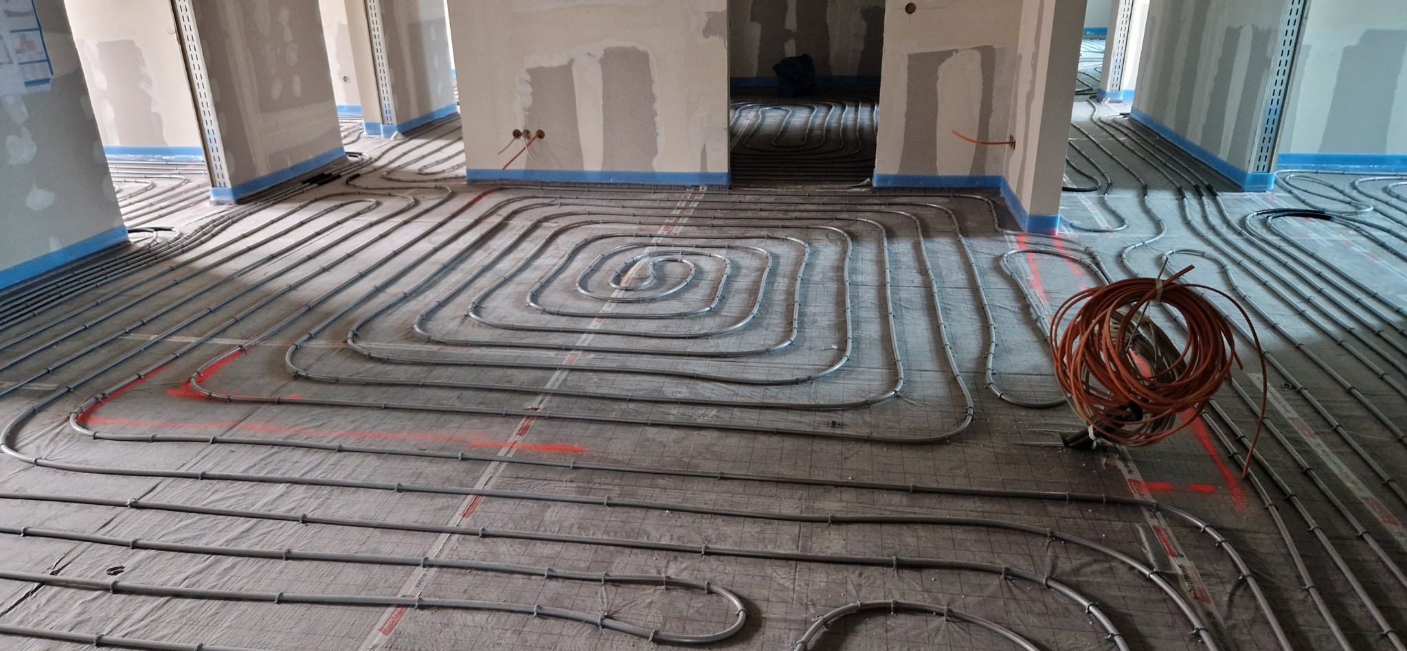 Szare rury ogrzewania podłogowego ułożone w spiralny wzór na podkładzie z siatki, widoczne także ściany w stanie surowym i zwój pomarańczowego kabla.