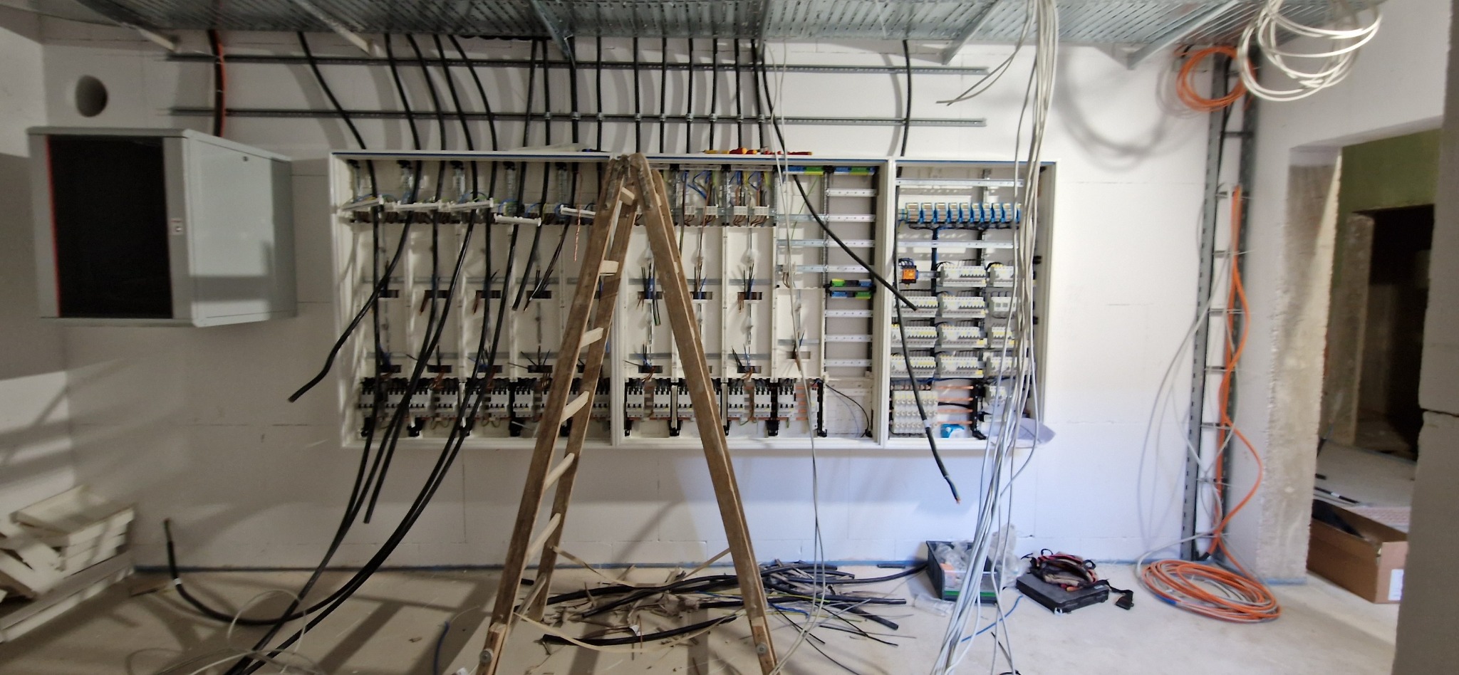 Szafa rozdzielcza z licznymi przewodami elektrycznymi w surowym wnętrzu, widoczna drewniana drabina i narzędzia.